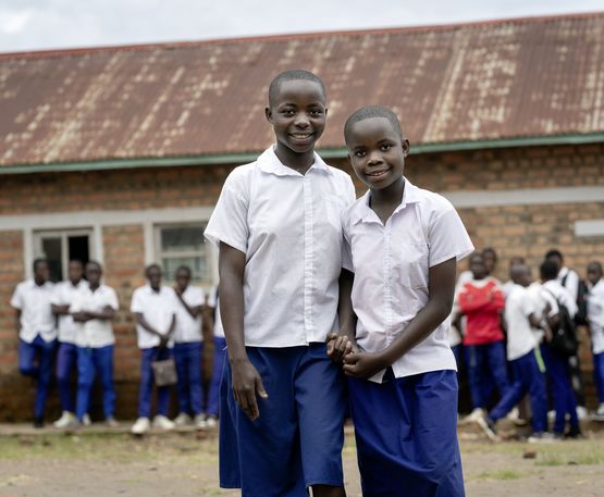 Die Schülerinnen Bien Aimé Ambire Namegabe und ihre kleinere Schwester Birugu Fortune Namegabe  auf dem Schulhof, andere Schüler*innen im Hintergrund