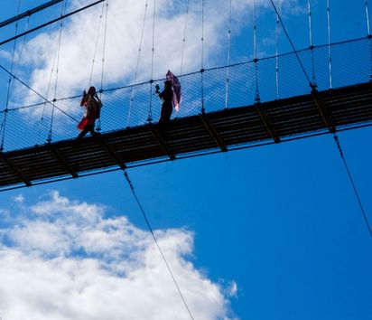 zwei Personen gehen über eine Hängebrücke vor blauem Himmel