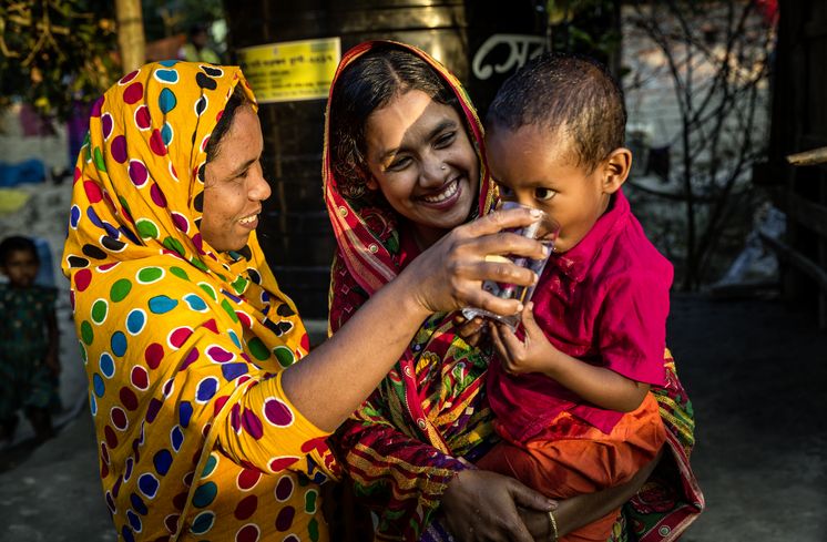 Sayra Begum (m.), profitiert von ihrer Regenwassergewinnung, die von CCDB gestellt wurde und kann jetzt sauberes Trinkwasser aus dem Wassertank zapfen und ihrem Sohn geben. Die Menschen in den kleinen Siedlungen an den Fluessen in der Naehe von Shyamnagar in Sued Bangladesch, spuehren deutlich den Klimawandel. Der Meeresspiegel steigt und Daemme werden zerstört. Da durch werden Felder und die wichtigen Suesswasserteiche ueberflutet und versalzen. CCDB Bangladesch arbeitet an Suesswassergewinnungs-Projekten und sorgt dafuer, dass die Menschen nach und nach sauberes Trinkwasser erhalten.
