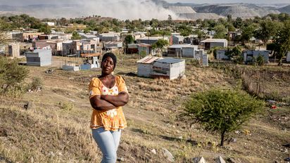 Porträt von Christina Mdau, im Hintergrund die Gemeinde hinter der Staub aus der Mine aufsteigt