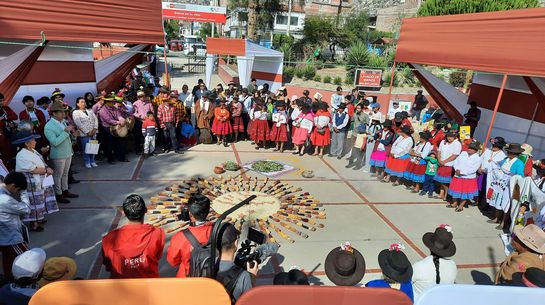 Eine Gruppe von Menschen in moderner und traditioneller indigener Kleidung umringt Saatgut das inmitten eines gepflasterten Platzes liegt..