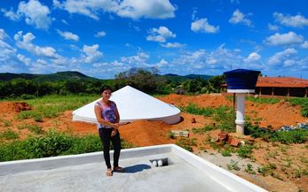 Frau steht auf einem gepflasterten Platz im Hintergrund eine Beton-Zisterne
