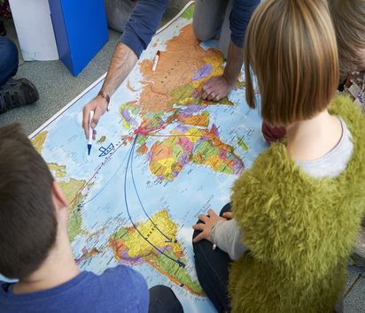 zwei Kinder sitzen vor einer Weltkarte auf die etwas eingezeichnet wird 
