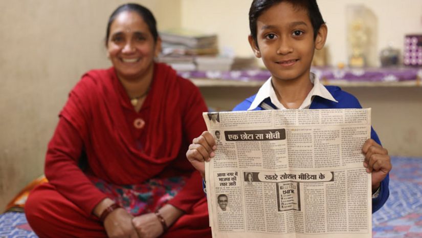 Ein Junge hält eine Zeitung in die Kamera, eine Frau sitzt im Hintergrund