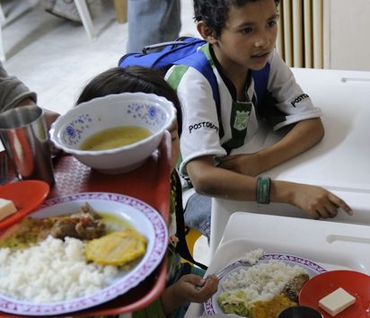 Kinder sitzen an einem Tisch in der Schulkantine. Auf den Tabletts haben sie ein reichhaltiges Essen.