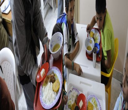 Kinder sitzen an einem Tisch in der Schulkantine. Auf den Tabletts haben sie ein reichhaltiges Essen.