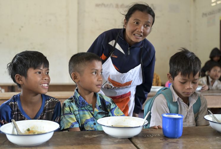 Drei Schüler sitzen in der Kantine vor ihren Tellern. Hinter ihnen steht ein Frau mit Schürze.